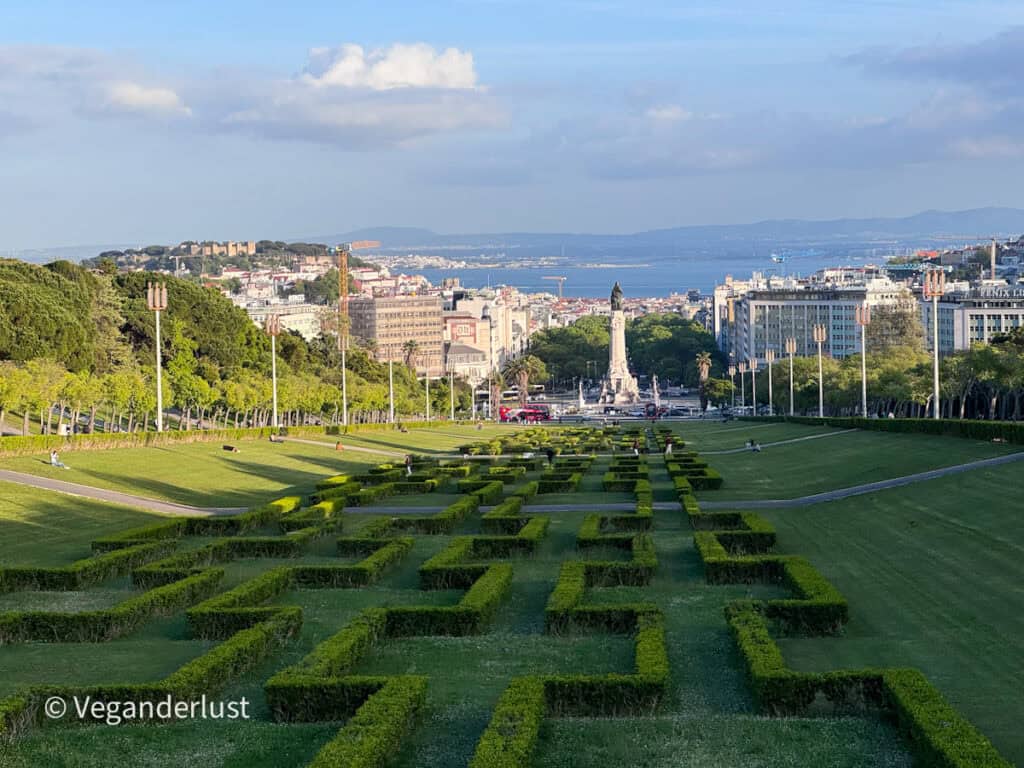 Formal garden with neatly trimmed hedge patterns leads down a wide green park toward a tall monument and city buildings with a river in the distance. People sit and stroll along the grassy slopes under a blue sky with scattered clouds.