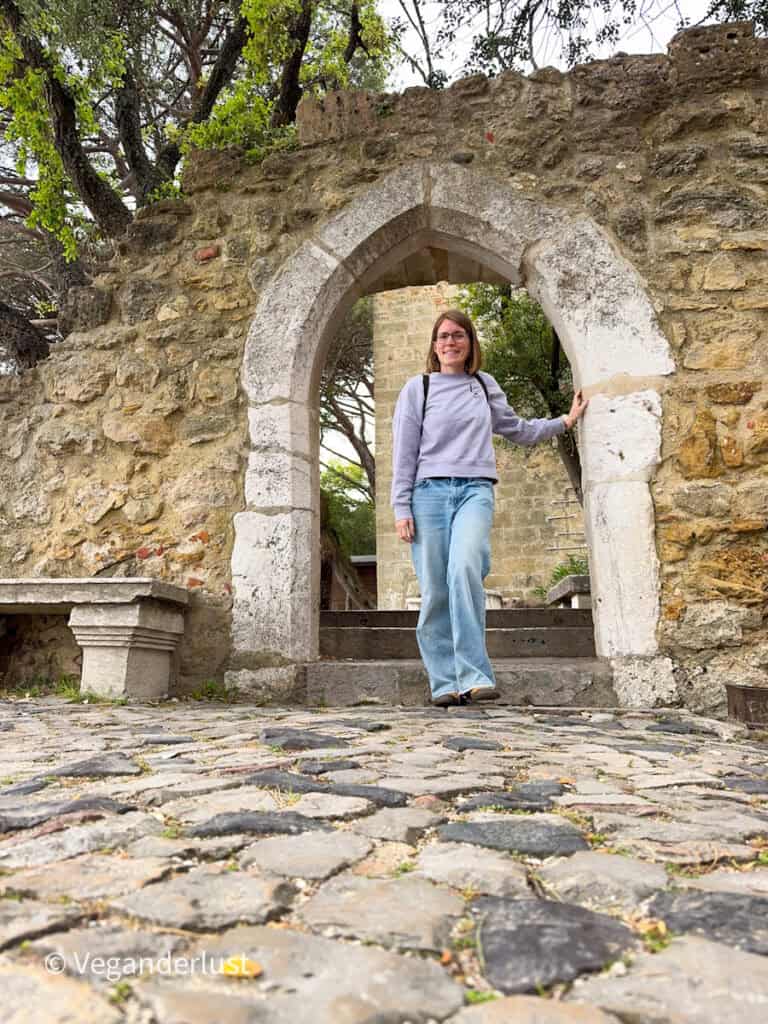 Tina wearing glasses walks through a small stone archway in a castle wall, stepping onto a cobblestone path with trees and historic ruins surrounding her.