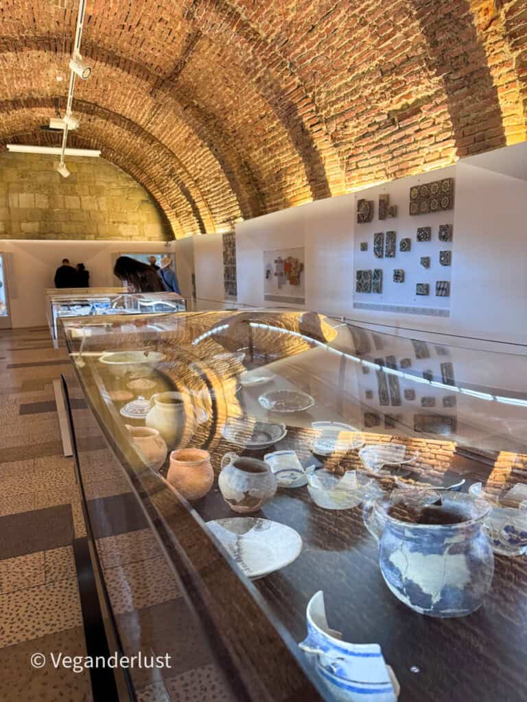 Museum interior with arched brick ceilings and glass display cases showcasing pottery and artifacts arranged along a long table.