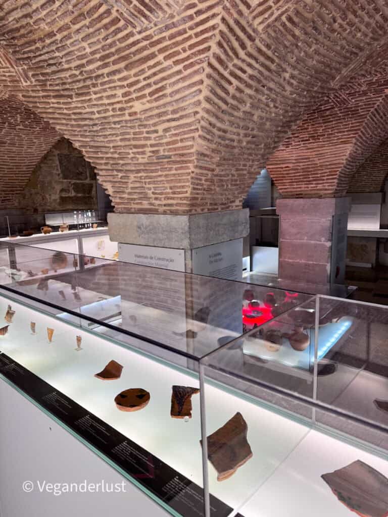 Close view of brick vaulted ceilings above glass cases displaying pottery fragments with a sign reading "Materiais de Construção Construction Material" and "A Cozinha The Kitchen".