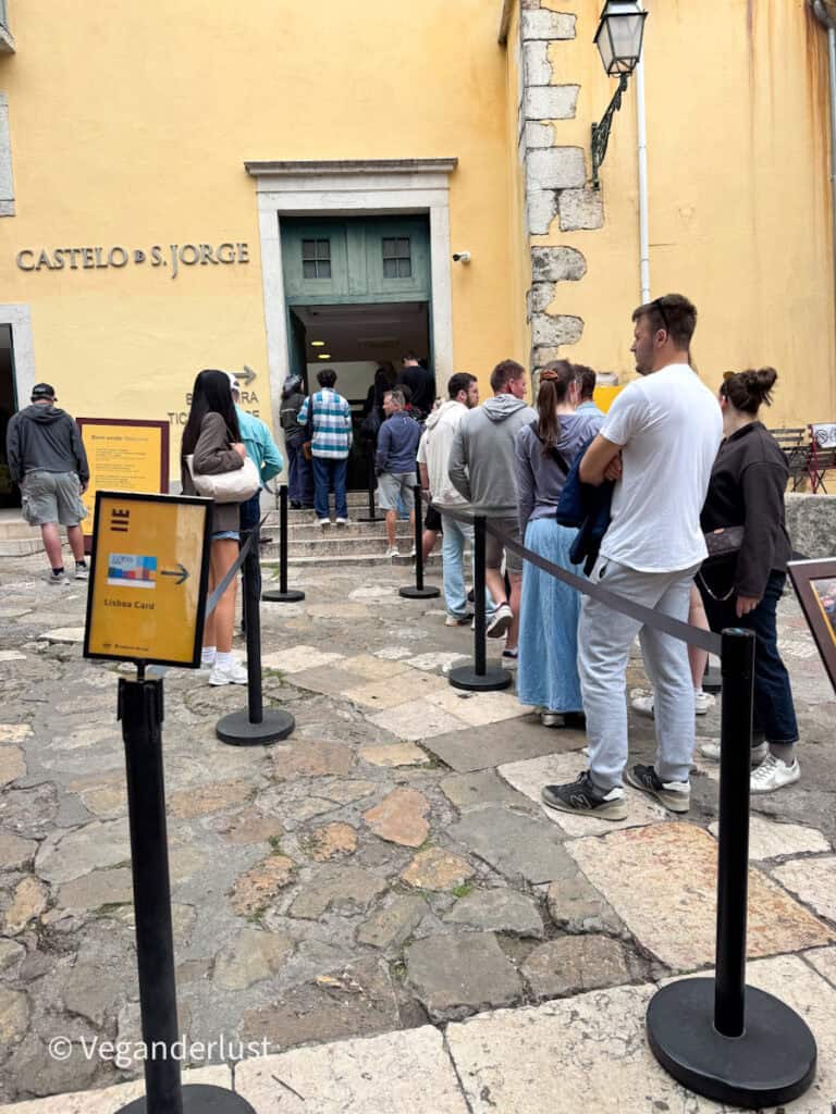 People stand in a queue outside a yellow building entrance with a sign reading "CASTELO DE S. JORGE" and a nearby sign displaying "Bilheteira Tickets Lisboa Card" with an arrow.