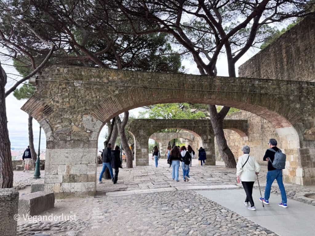 Stone walkway lined with arches and trees where visitors stroll along a cobblestone path through the castle grounds.