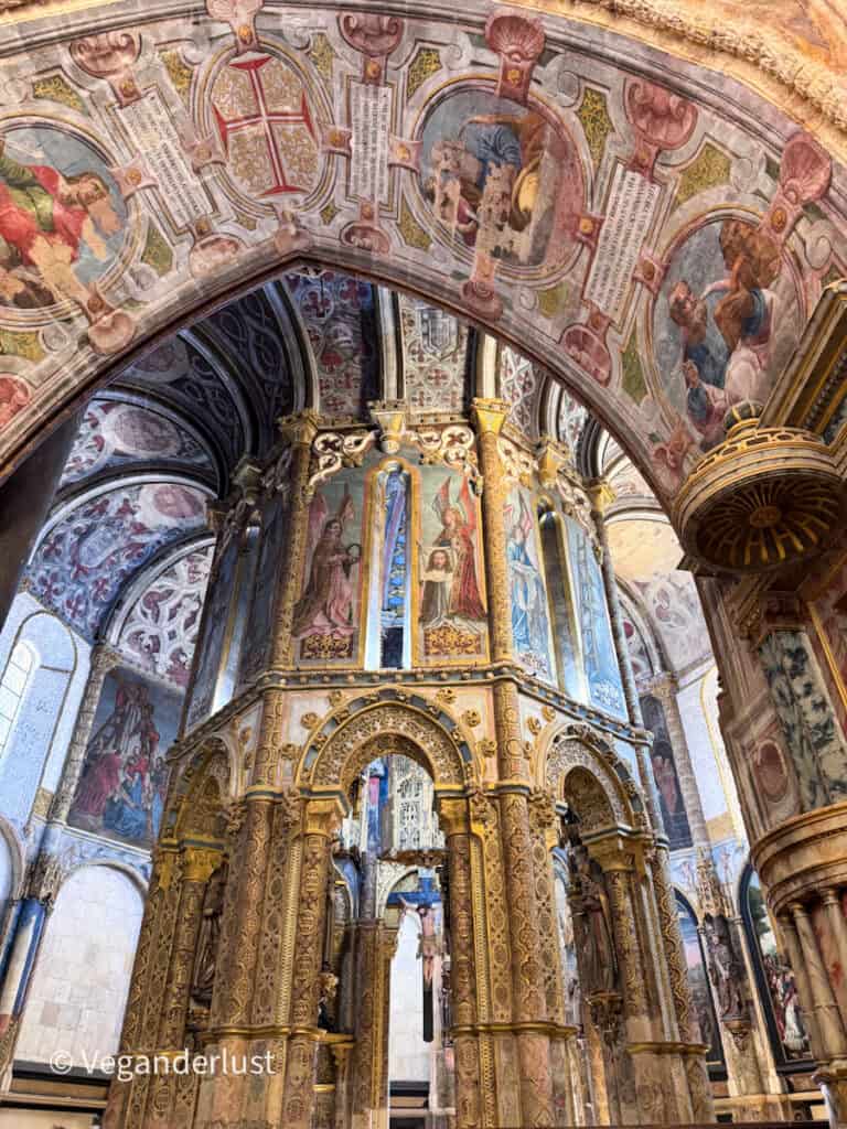 Ornate church interior with a richly decorated golden structure covered in religious paintings and carvings beneath a painted arch ceiling. A crucifix hangs at the center surrounded by detailed columns and frescoes.