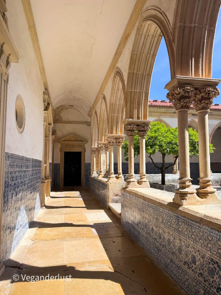 Sunlit cloister walkway with stone arches and columns casting shadows along a tiled floor. Blue and white patterned tiles line the walls and a small tree stands in the courtyard.
