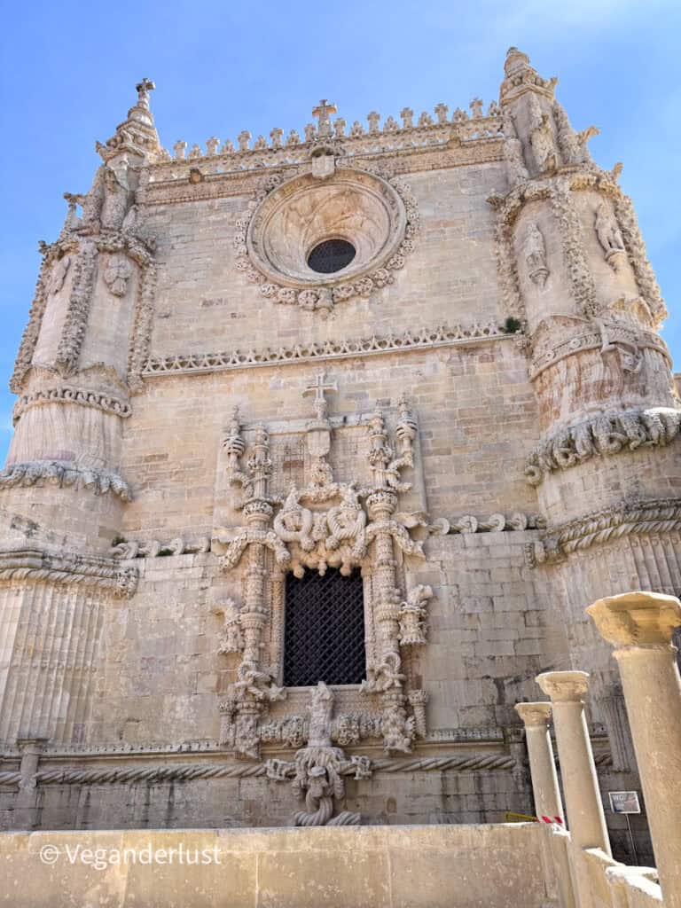 Highly detailed stone church facade with intricate carvings, statues, and a circular window at the center. Twisted rope like stonework and decorative elements frame the window and doorway against a clear blue sky.