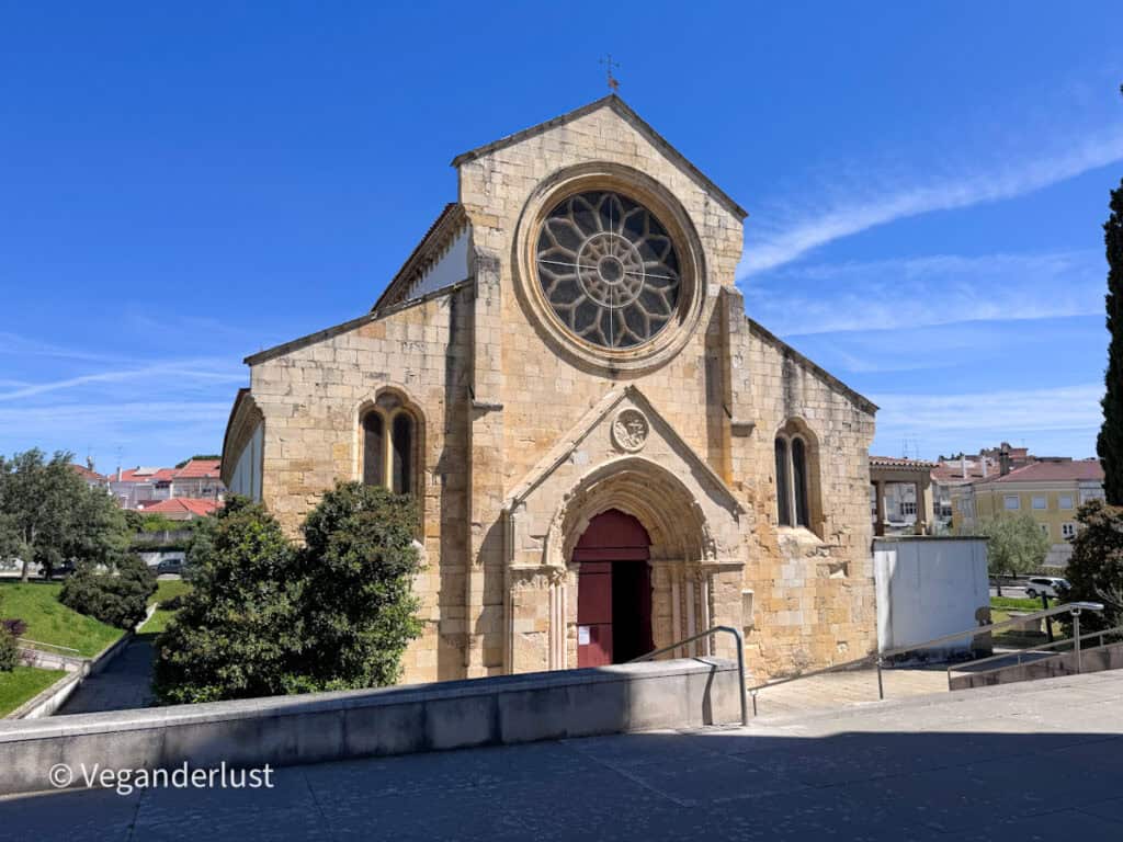 Small stone church with a large circular rose window above a pointed doorway. The building sits under a clear blue sky with nearby houses in the background.