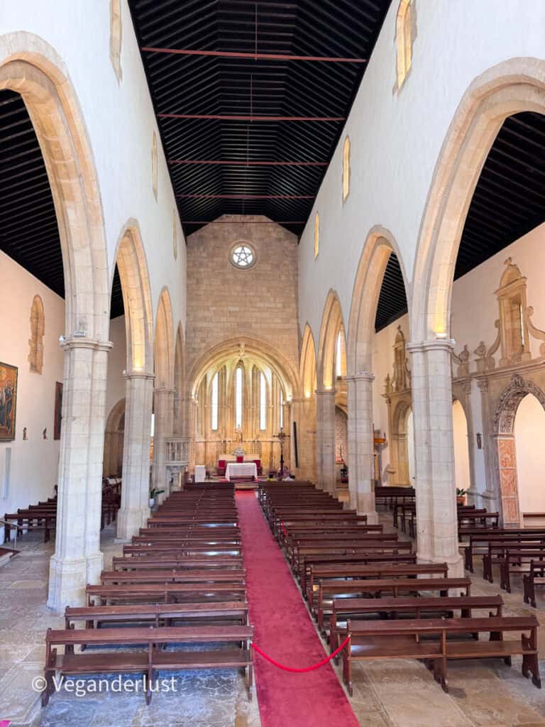 Interior of a historic church with stone arches, wooden pews, and a red carpet aisle leading to the altar. Soft light filters in from high windows along the walls.