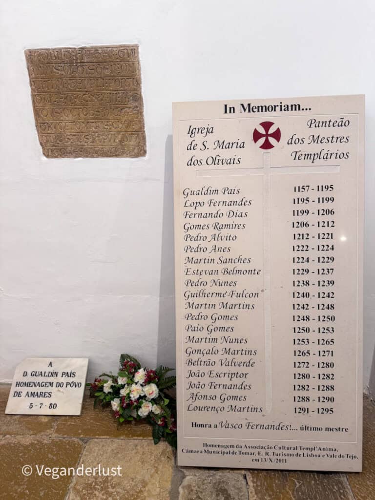 Memorial plaques mounted on a white wall inside a church with engraved names and dates. Flowers rest at the base beside a smaller stone inscription.
