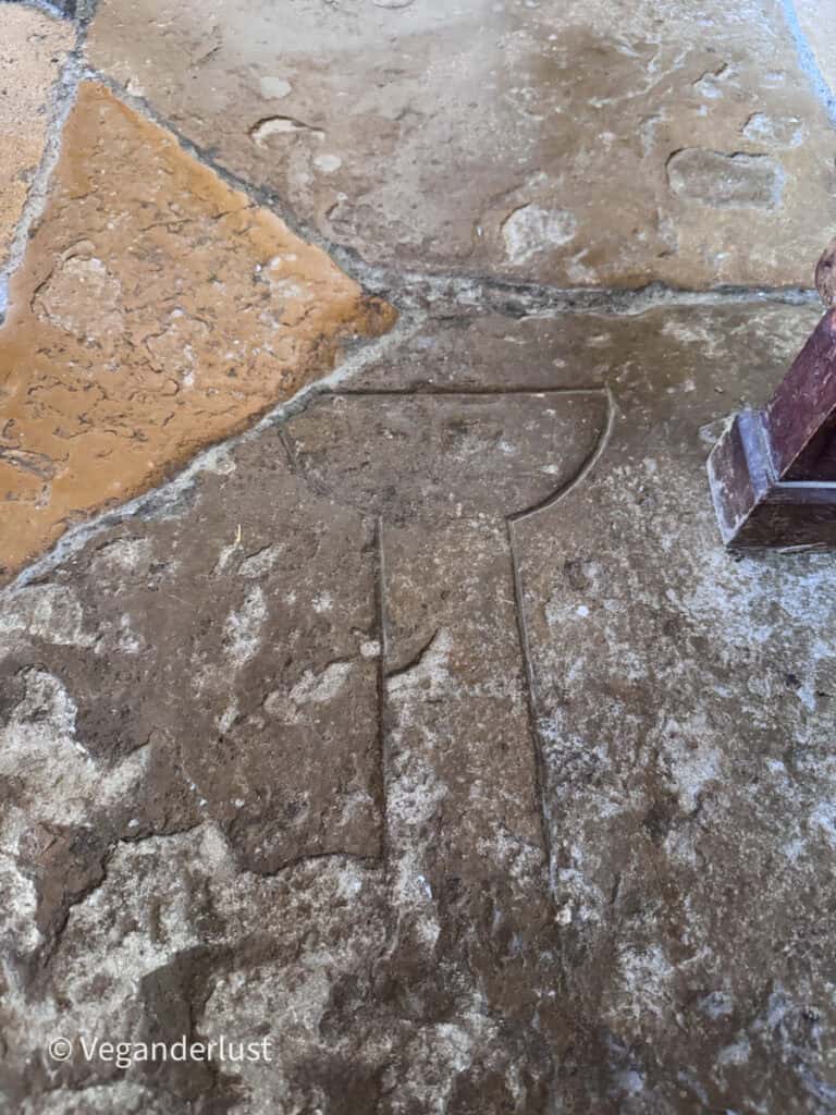 Worn stone floor inside a building with a carved outline resembling a chalice shape. The texture of the aged stone is rough and uneven.