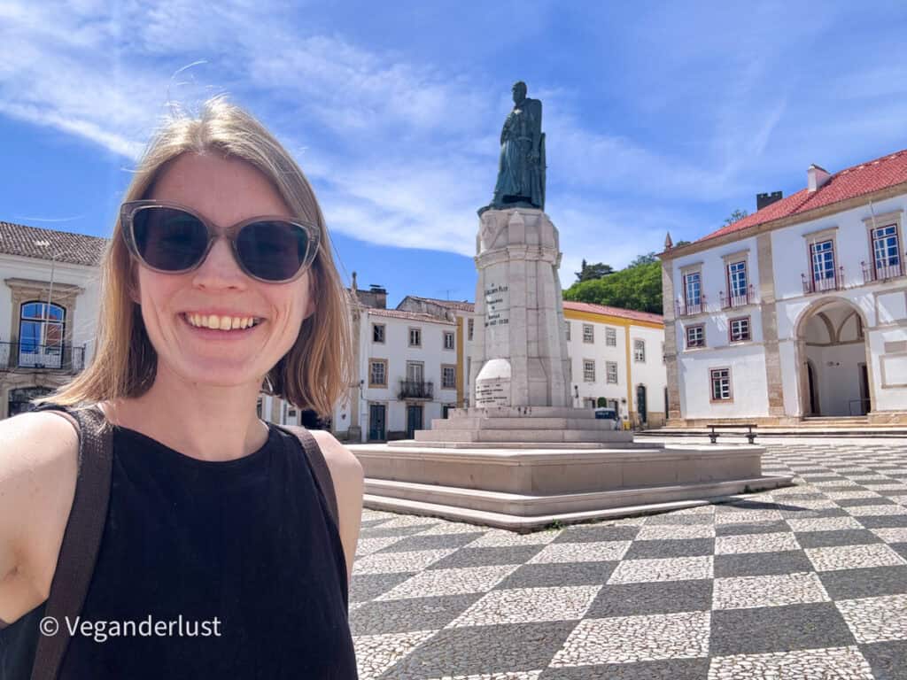 Tina wearing sunglasses smiles at the camera in a sunlit town square with a patterned stone floor. A statue on a pedestal and historic buildings with red tiled roofs stand behind her under a blue sky.
