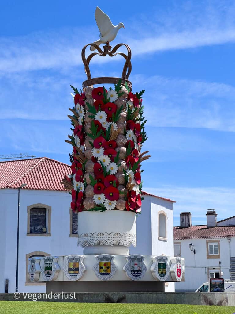 Decorative monument shaped like a tall column covered in red and white flowers and wheat motifs with a white dove on top. A row of crests decorates the base in front of white buildings with tiled roofs.
