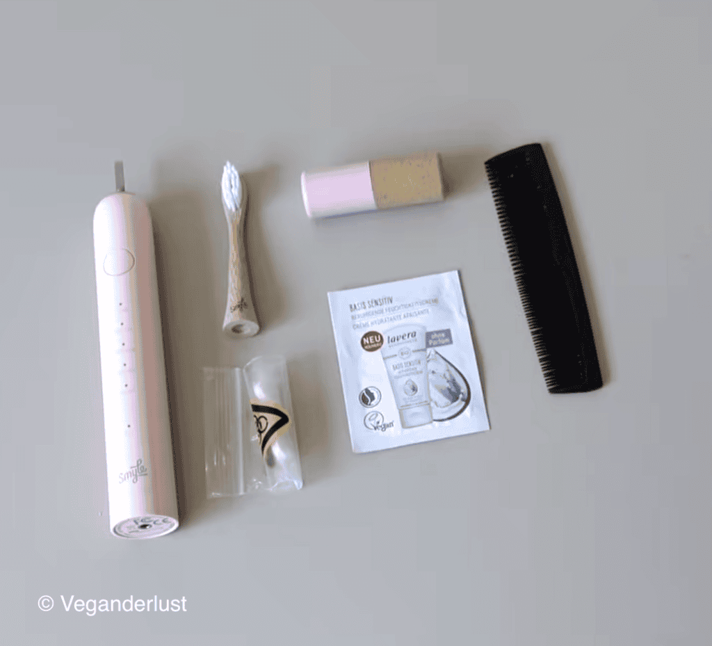Small personal care items arranged on a light surface including a pink electric toothbrush, a bamboo toothbrush, a pink lip balm tube, a black comb, a clear plastic bag with earplugs, and a skincare sample packet labeled "BASIS SENSITIV" and "lavera" with "NEU" and "Vegan" visible. The items are spaced out to show a compact hygiene kit for travel or daily use.