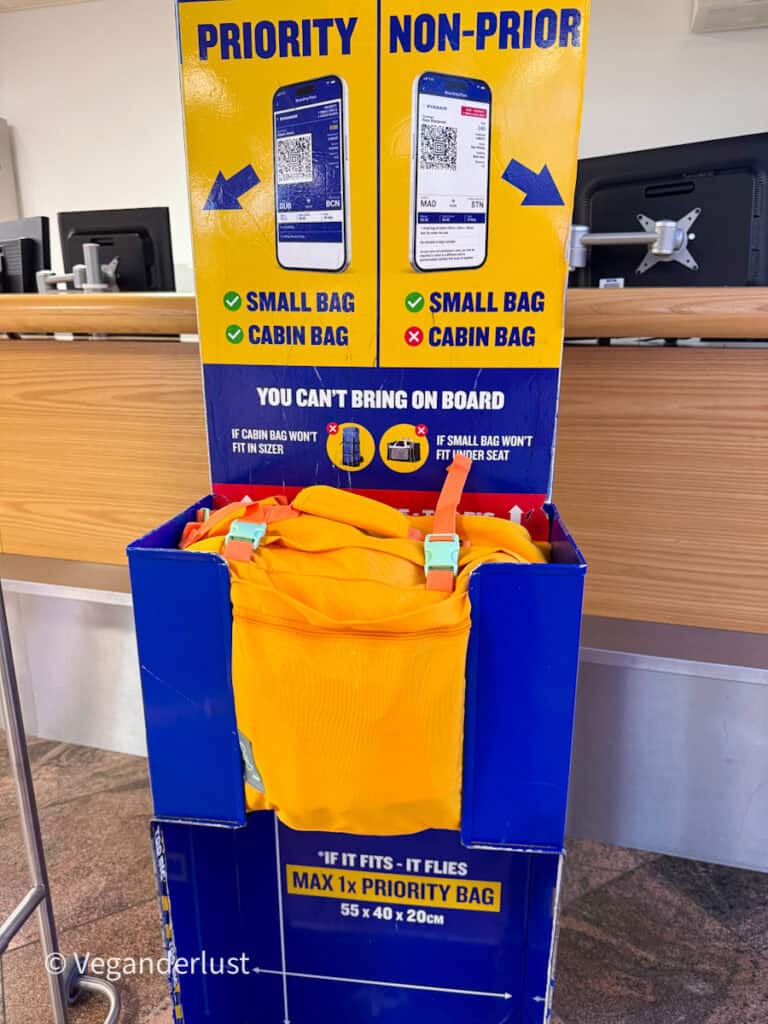 Bright yellow backpack placed inside an airline cabin bag size checker at an airport check in counter. Signs above the bag compare “PRIORITY” and “NON PRIORITY” boarding allowances and show that priority passengers can bring both a small bag and cabin bag while non priority passengers can only bring a small bag. Text on the size checker reads “YOU CAN’T BRING ON BOARD IF CABIN BAG WON’T FIT IN SIZER” and “MAX 1x PRIORITY BAG 55 x 40 x 20CM.”