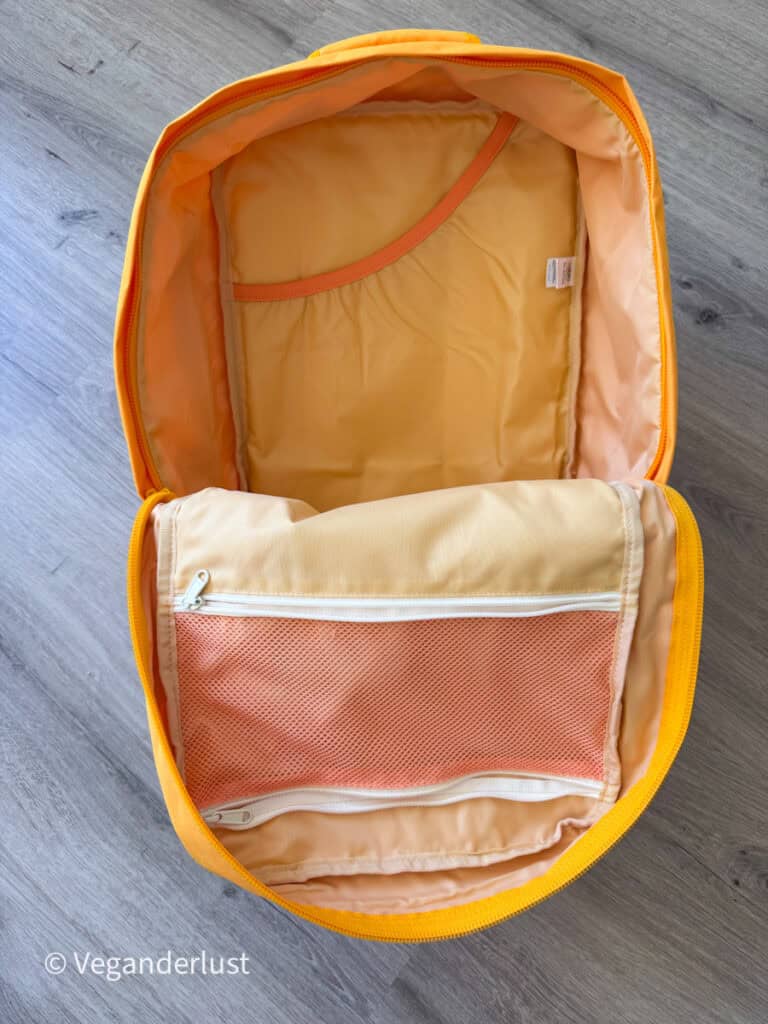 Open bright yellow backpack lying on a wooden floor with the main compartment unzipped to show the interior storage space. The bag has a large mesh zippered divider pocket and a padded sleeve pocket inside.