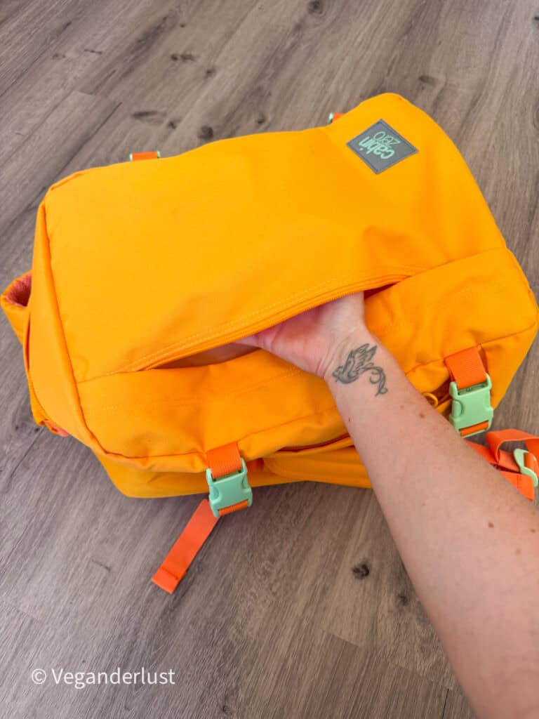 Close up of a bright yellow backpack on a wooden floor with a hand sliding into a hidden zippered pocket near the back panel. Orange straps and mint green buckles are visible on the outside of the bag.