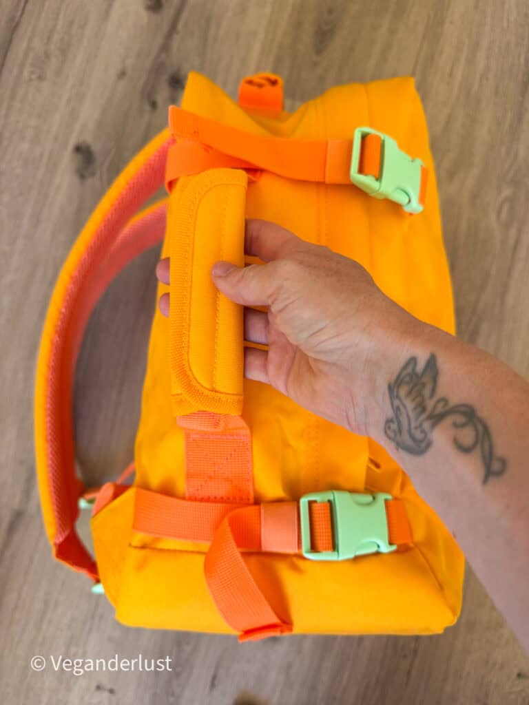 Side view of a bright yellow backpack showing the padded grab handle on the top of the bag. A hand holds the handle while orange compression straps and mint green buckles wrap around the backpack.