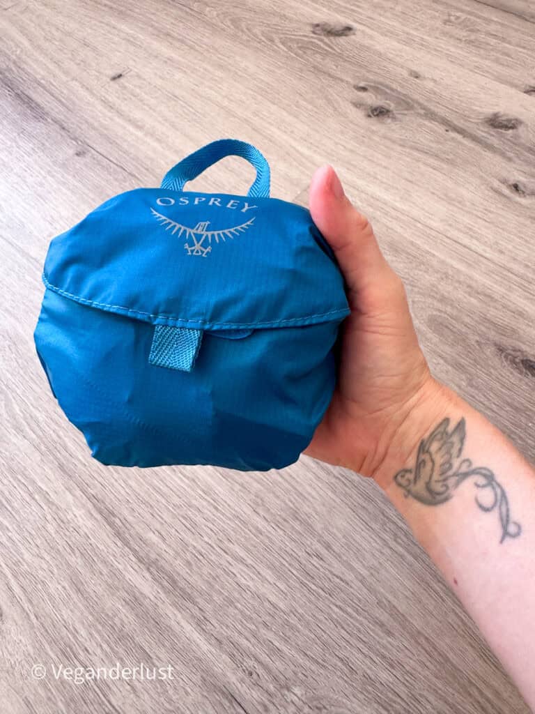 Hand holding a compact blue Osprey foldable backpack packed into a small pouch with a carry loop on top. The lightweight backpack is folded down to fit in one hand.