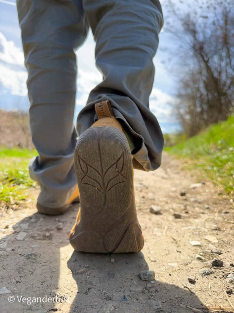 Close up of the sole of a mustard yellow walking shoe as a person hikes along a dirt trail. The shoe is lifted mid step with grass, trees, and a bright sky blurred in the background.