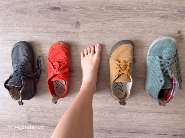 Bare foot positioned between four different barefoot shoe styles arranged on a wooden floor. The comparison shows how the natural shape of the foot aligns with the wide toe boxes of the shoes, which are displayed in black, coral red, mustard yellow, and light green colors.