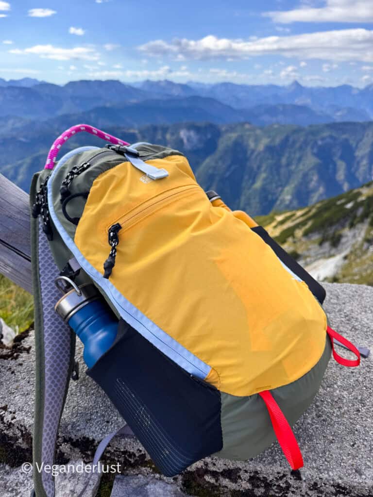 Yellow and green hiking backpack with a blue water bottle resting on a rocky mountain viewpoint. Layered mountain ridges stretch into the distance beneath a blue sky.