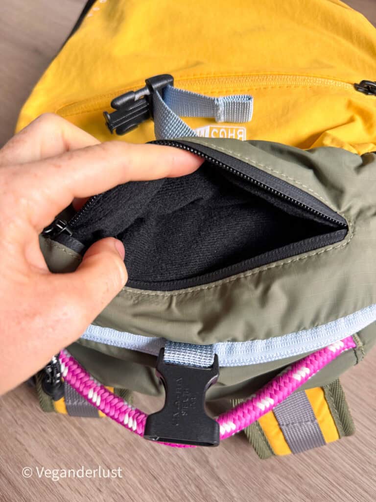 Hand holds open a small zippered pocket on the green front section of a yellow and green backpack. The pocket has a soft black lining with a gray buckle strap and bright pink cord visible below.