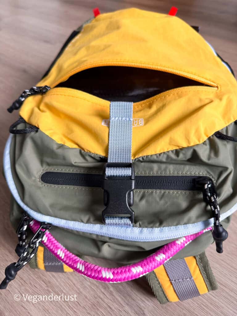 Front of a yellow and green backpack showing an open top zippered compartment. A gray buckle strap crosses the center with another zippered pocket and bright pink cord below.